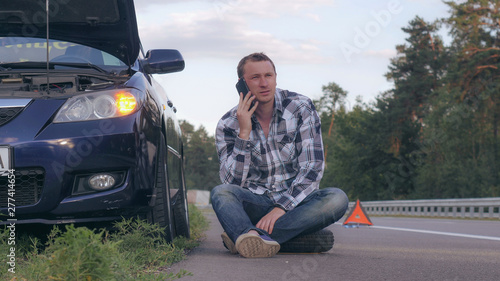 Εκτύπωση καμβά Caucasian young man sitting on the road nearby broken car talking using smartphone