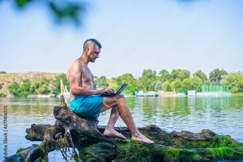 Freelancer working on a laptop while sitting on a fallen tree near the river. Freelancer working on a laptop while sitting on a fallen tree near the river.