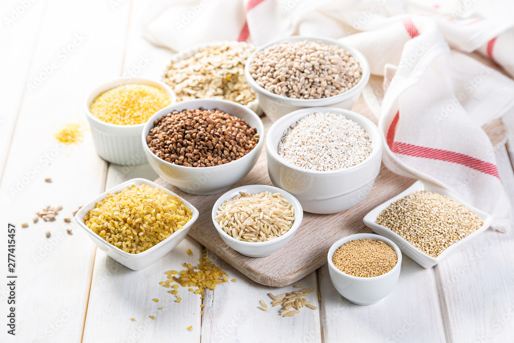 Selection of whole grains in white bowls - rice, oats, buckwheat, bulgur, porridge, barley, quinoa, amaranth, on white wood background