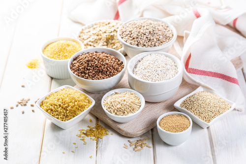 Selection of whole grains in white bowls - rice, oats, buckwheat, bulgur, porridge, barley, quinoa, amaranth, on white wood background photo