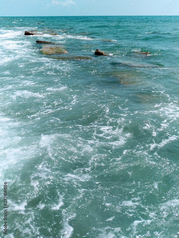 Naklejka premium abstract background of bright turquoise sea water, under the water a huge concrete stones as the breakers, the white foam of the surf, the sunlight sparkle on the ocean waves, a strip of azure sky