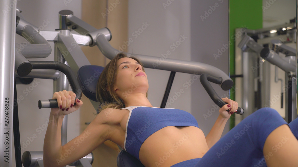 Woman doing exercises on biceps training machine in gym. Caucasian bodybuilder trains alone. Young beautiful woman working out lifting weights. Girls wearing in blue sport top. Woman doing exercises on biceps training machine in gym. Caucasian bodybuilder trains alone. Young beautiful woman working out lifting weights. Girls wearing in blue sport top.