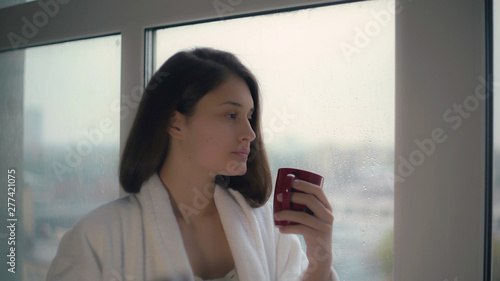 Portrait young woman standing near window holding red cup. Autumn season with rain. Thoughtful melancholy lady with brown hair wrapping up in white bathrobe.