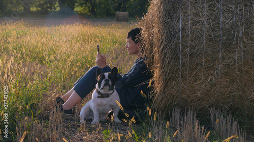 girl with dog resting messaging on smartphone outdoors. Attractive female wearing overalls texting message on mobile. Brunette enjoy nature and fresh air at field in summer season © vadosloginov