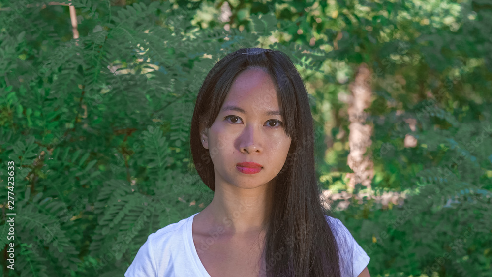 portrait asian woman with friendly smile in the park. Happy young girl standing in front of green tree outdoors. Vietnamese female wearing in casual t-shirt smiling looking at the camera. Her long portrait asian woman with friendly smile in the park. Happy young girl standing in front of green tree outdoors. Vietnamese female wearing in casual t-shirt smiling looking at the camera. Her long