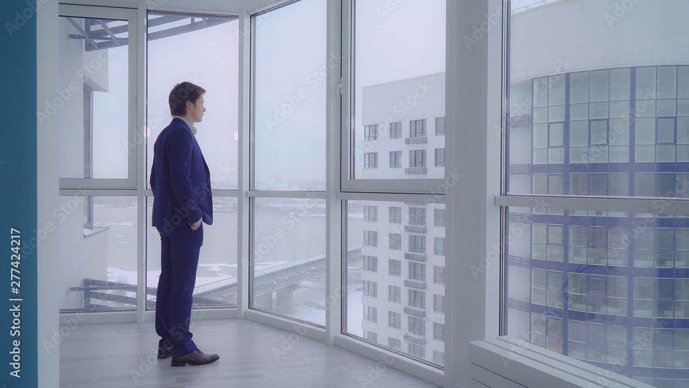 businessman in empty room with big panoramic windows looking on the urban city view. young professional man wearing in elegant suit alone in modern apartment. caucasian male deep in thought. businessman in empty room with big panoramic windows looking on the urban city view. young professional man wearing in elegant suit alone in modern apartment. caucasian male deep in thought.