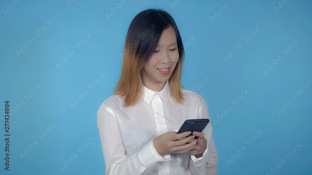 young asian woman messaging on mobile with touch screen on blue background in studio. attractive millennial girl wearing white casual shirt looking at the camera smiling young asian woman messaging on mobile with touch screen on blue background in studio. attractive millennial girl wearing white casual shirt looking at the camera smiling