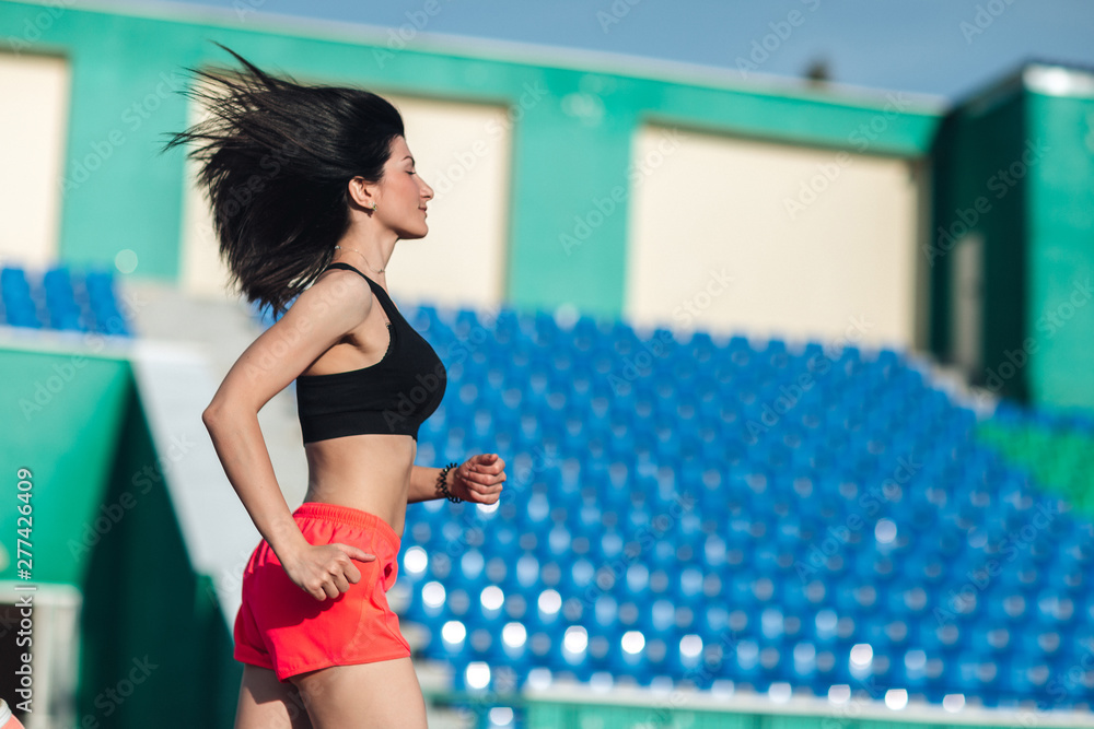 Girl running track on stadium. Real side view of young brunette woman in pink shorts and tank top and pink sneakers. Outdoors, sport