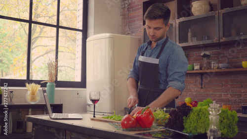 Young man cooking on the kitchen. Handsome guy cutting salad at home wearing in casual shirt and apron. Modern interior with beautiful view through the big window. On cook table laptop and wineglass.
