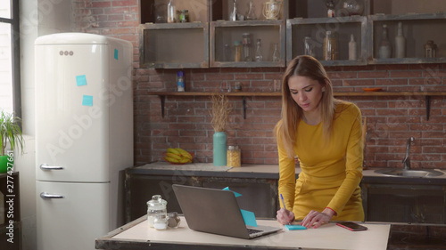 businesswoman writing notes in kitchen. Adult woman working at home on the cooking table laptop. Caucasian lady wearing in elegant dress glues blue sticky notes on fridge in apartmen