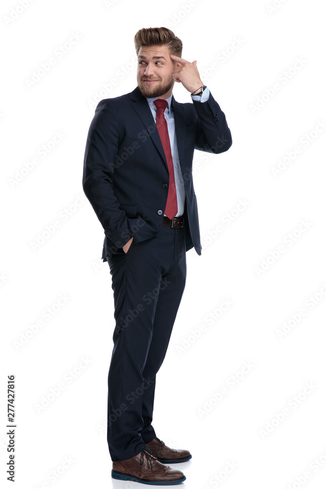 Young businessman pointing to his forehead and holding his hand in his pocket while looking over his shoulder and wearing a blue suit, standing on white studio background Young businessman pointing to his forehead and holding his hand in his pocket while looking over his shoulder and wearing a blue suit, standing on white studio background