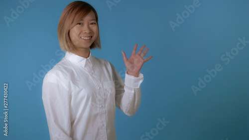 portrait young asian female greeting waving hand on blue background in studio. attractive korean woman with blond hair wearing white casual shirt looking at the camera with smile.