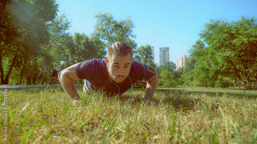 A handsome man in trendy outfit makes push ups during the video. He is wearing a blue T-shirt and an armband for the smartphone. The male is working out in the park with an urban background. A handsome man in trendy outfit makes push ups during the video. He is wearing a blue T-shirt and an armband for the smartphone. The male is working out in the park with an urban background.