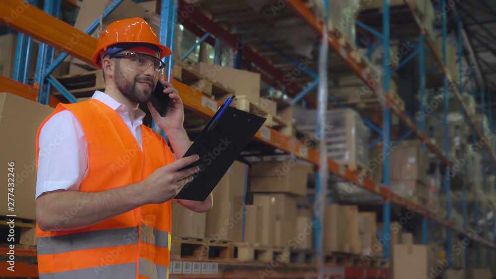 Portrait manager at work in warehouse has phone conversation with client. Handsome worker talking by smartphone discussing the logistics. Man wearing hard hat and orange vest Portrait manager at work in warehouse has phone conversation with client. Handsome worker talking by smartphone discussing the logistics. Man wearing hard hat and orange vest