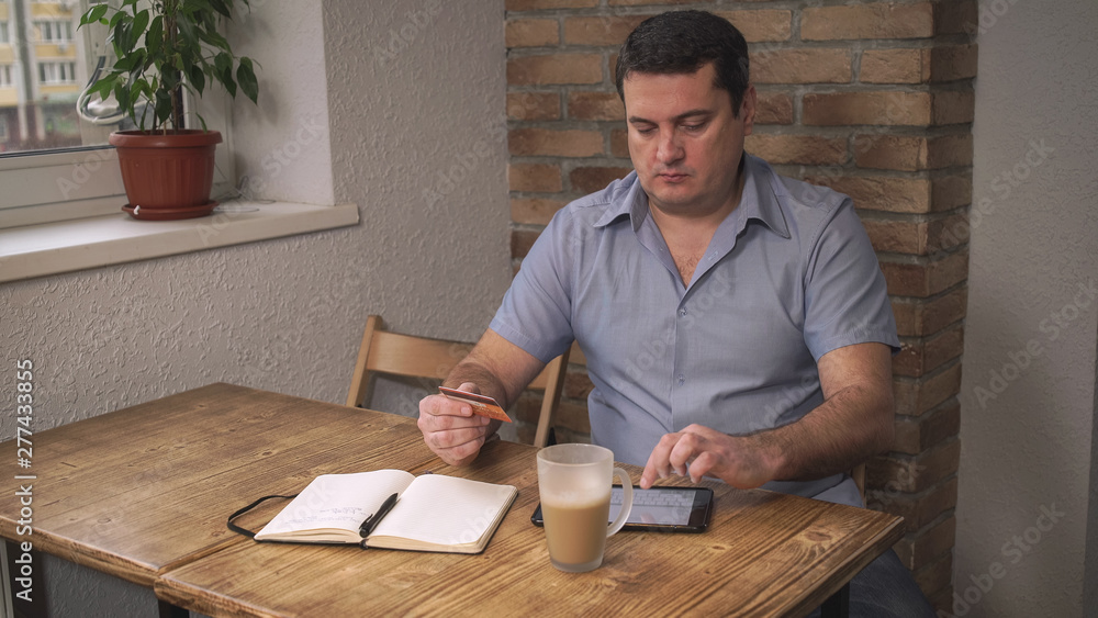 The man sitting in the cafe, pay for purchases online with credit card. On the table is a coffee and a notebook lies. Soft panorama The man sitting in the cafe, pay for purchases online with credit card. On the table is a coffee and a notebook lies. Soft panorama