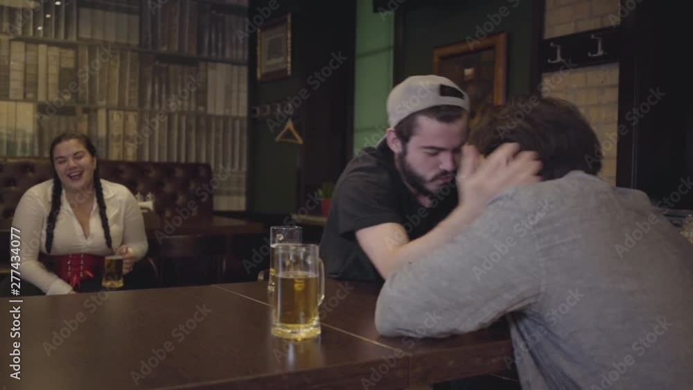 Two men sitting at a table in a bar, engaged in arm wrestling. Plump woman sitting in the background, emotionally supporting friends. Guys having fun together drinking beer. Leisure in a beer pub Two men sitting at a table in a bar, engaged in arm wrestling. Plump woman sitting in the background, emotionally supporting friends. Guys having fun together drinking beer. Leisure in a beer pub