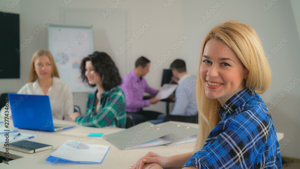 Fototapeta premium Portrait attractive woman with long blond hair in office startup company. Young businesswoman looking at the camera with candid smile. Professional female with natural makeup smiling. On the