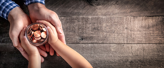 Hands Of Father Giving Jar Of Coins To Child On Wooden Table Background - Inheritance / Parent Providing For Children Concept