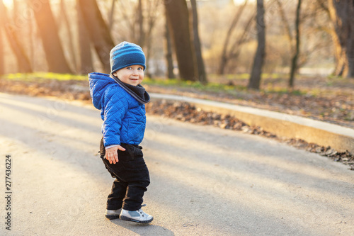 Portrait of cute funny caucasian toddler boy in blue jacket and hat enjoying walking at autumn park or forest during sunset with sun rays shining through trees on background. Baby having fun outdoors