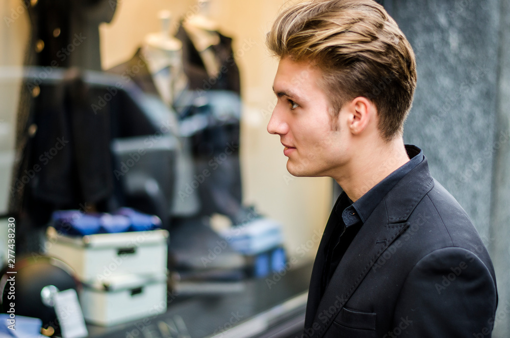 Handsome Young Man in Black Elegant Suit Looking at Displayed Fashion Items in Glass Window Boutique at the Street Side. Handsome Young Man in Black Elegant Suit Looking at Displayed Fashion Items in Glass Window Boutique at the Street Side.