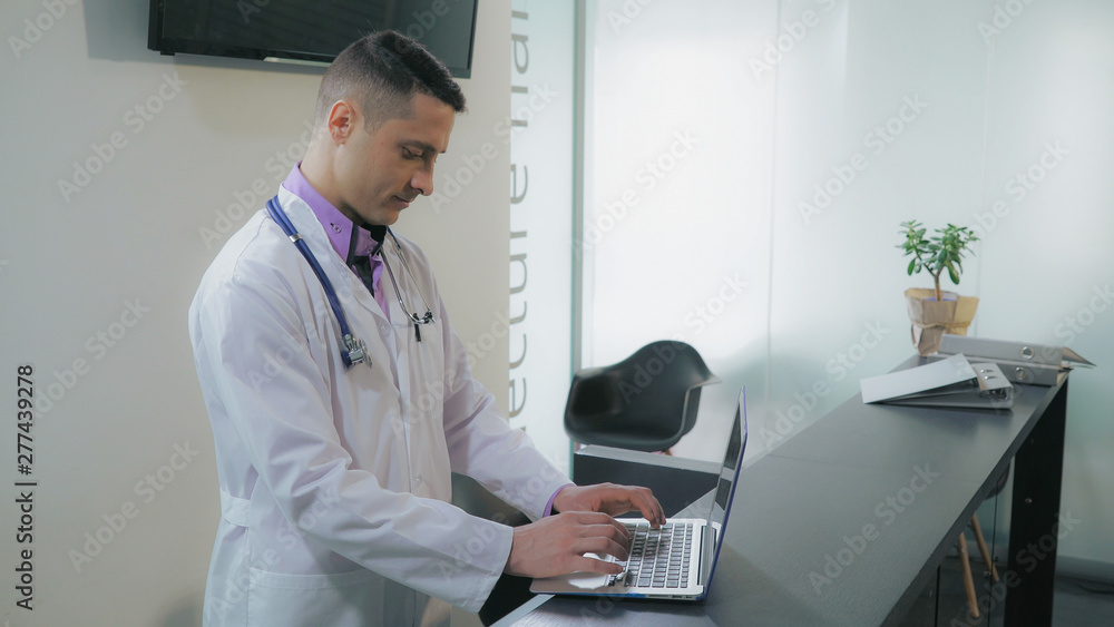 Handsome doctor or nurse using computer looking on screen laptop typing on keyboard. Man assistant therapist standing at reception desk wearing in white coat. Handsome doctor or nurse using computer looking on screen laptop typing on keyboard. Man assistant therapist standing at reception desk wearing in white coat.