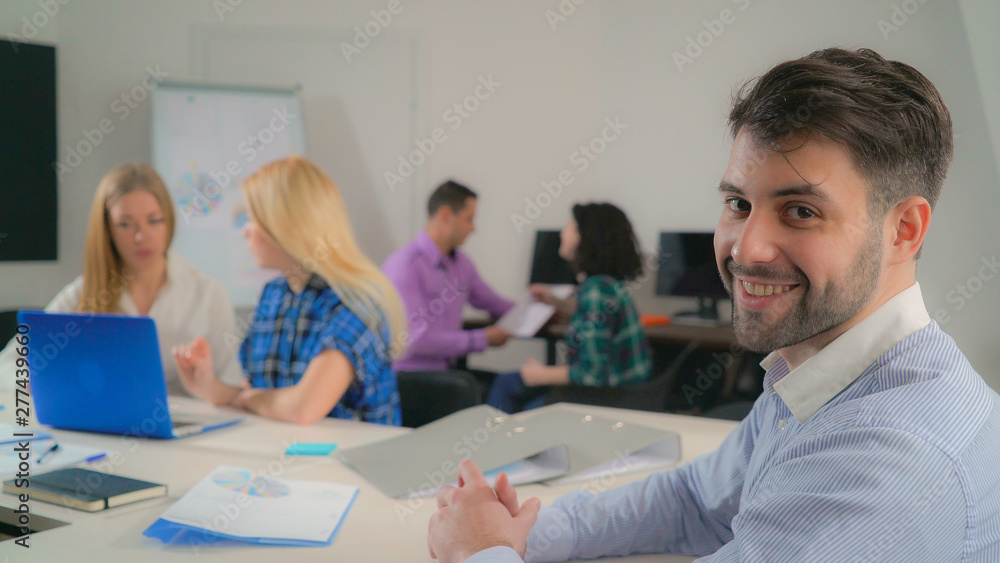 Portrait handsome mixed race man in office startup company. Young businessman looking at the camera with friendly smile. Professional male with beard smiling. On the background working business team. Portrait handsome mixed race man in office startup company. Young businessman looking at the camera with friendly smile. Professional male with beard smiling. On the background working business team.