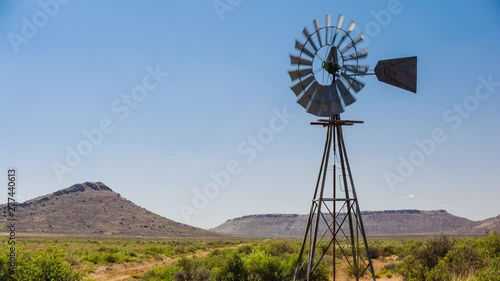 Loop of A static timelapse of a typical Karoo landscape scene filled with shrubs and grass with a windmill blowing frantically in the wind against a bright blue sky and hills in the background available on request.