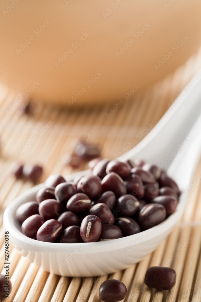 Raw, uncooked, dried adzuki (red mung) beans in white spoon on bamboo mat background Raw, uncooked, dried adzuki (red mung) beans in white spoon on bamboo mat background