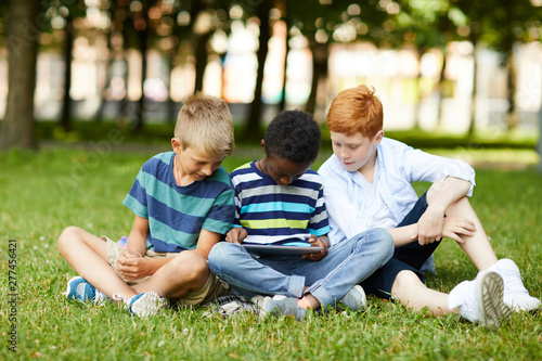 Three smiling teenage school friends sitting in park on grass and playing new tablet game together