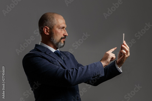 Bald middle-aged man with a beard, in a jacket with a white shirt and blue tie holding a mobile phone on a dirty gray background