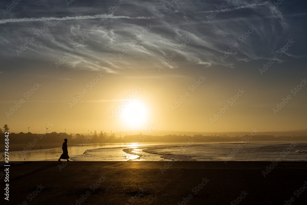 Man silhouette walking on sand beach at low tide at sunset with beautiful clouds in the background