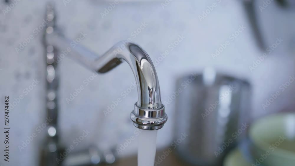 Clean tap water for washing dishes starts flowing from long, cute, new bright, steel tap in light kitchen when someone turned it on in daylight. Clean tap water for washing dishes starts flowing from long, cute, new bright, steel tap in light kitchen when someone turned it on in daylight.