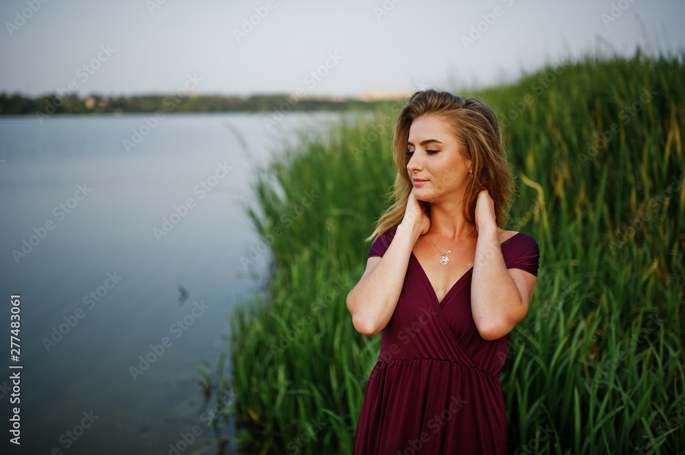 Blonde sensual woman in red marsala dress posing against lake with reeds. Blonde sensual woman in red marsala dress posing against lake with reeds.