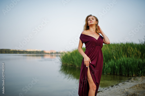 Blonde sensual woman in red marsala dress posing against lake with reeds.