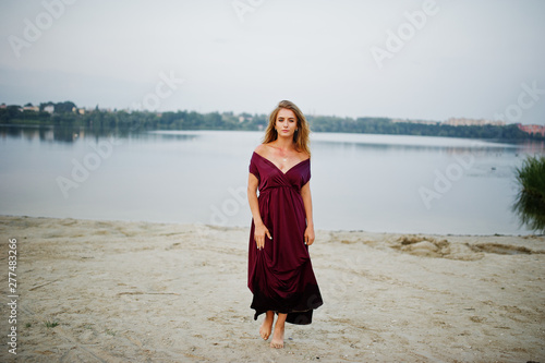 Blonde sensual barefoot woman in red marsala dress posing against lake on sand.
