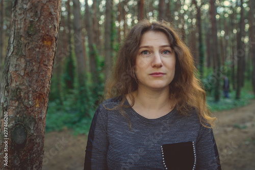 Serious woman looking out from behind tree. Pretty calm woman touching tree and looking at camera in quiet forest