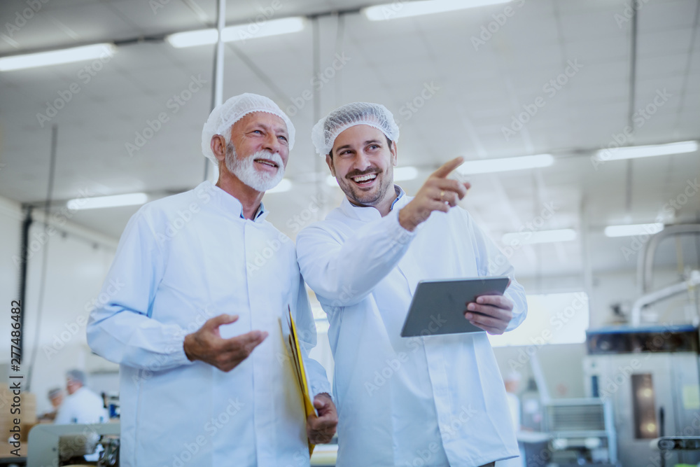 Two smiling Caucasian supervisors in white sterile uniforms standing in food factory. Younger one holding tablet and pointing while older one holding folder with documents under armpit. Two smiling Caucasian supervisors in white sterile uniforms standing in food factory. Younger one holding tablet and pointing while older one holding folder with documents under armpit.