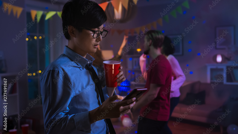 At the Wild House Party: Confident Asian Man Uses Smartphone Instead of Dancing With Other People.