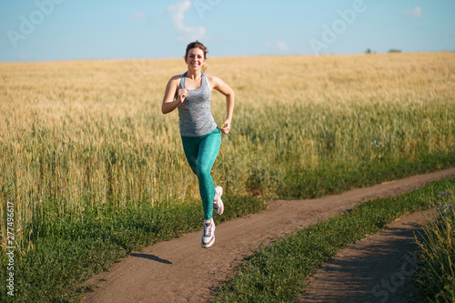 Sport, fitness, health care, cardio training. Active woman with athletic body running in the countryside. Smiling female jogger working out in the morning sunny day.
