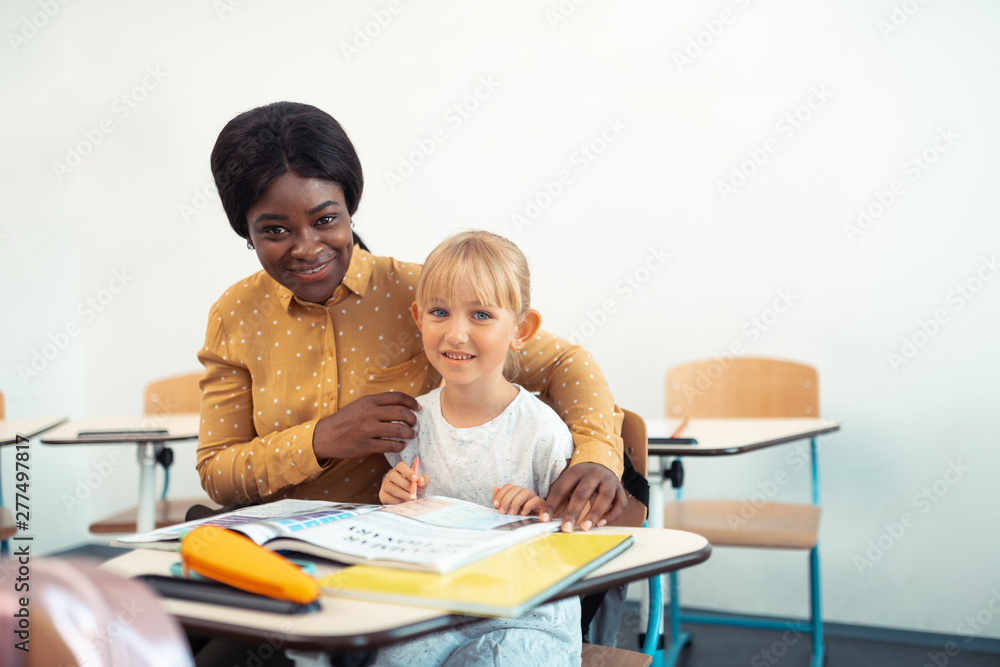 Beaming helpful teacher hugging cute blue-eyed smart girl Beaming helpful teacher hugging cute blue-eyed smart girl