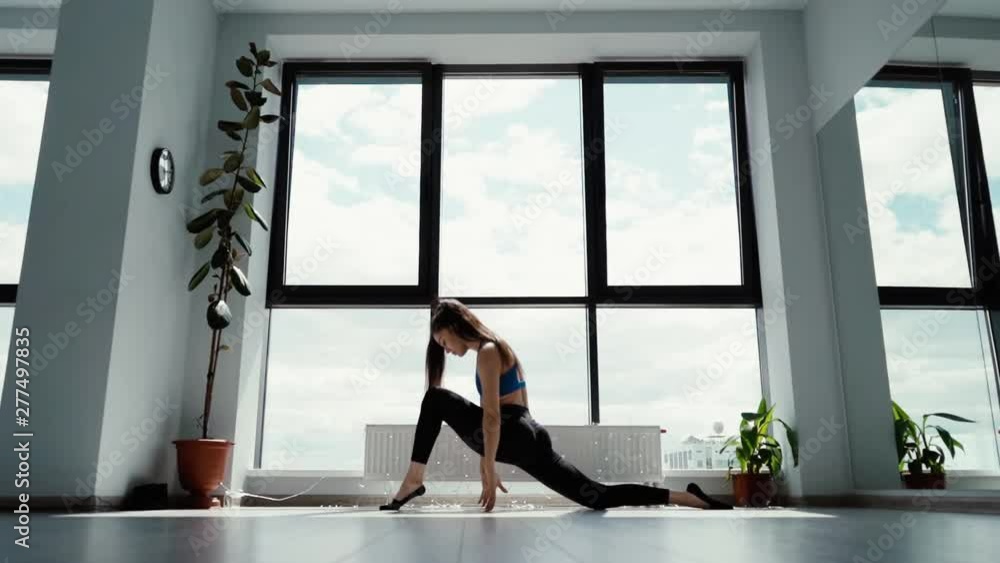 Flexible beautiful girl practices yoga and performs crescent asana in slow motion in light pilates class on background of large window. Young sportswoman makes low forward fall pose of Anjaneyasana. Flexible beautiful girl practices yoga and performs crescent asana in slow motion in light pilates class on background of large window. Young sportswoman makes low forward fall pose of Anjaneyasana.