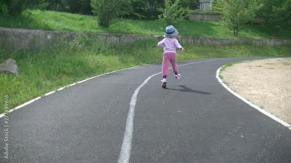 Little girl with blond hair and in a blue hat runs amusingly on a sports treadmill. Jumps up and waves. Summer. Back view. Little girl with blond hair and in a blue hat runs amusingly on a sports treadmill. Jumps up and waves. Summer. Back view.