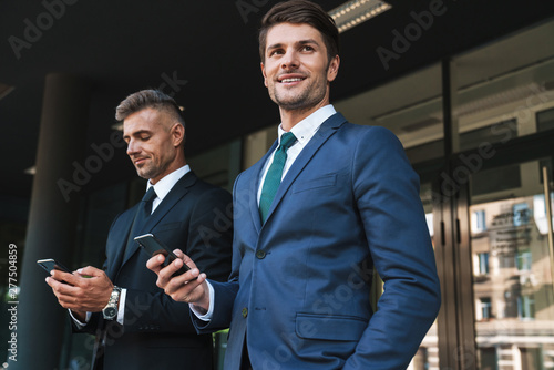 Image of joyful businessmen partners standing outside office center and holding cellphones together during working meeting