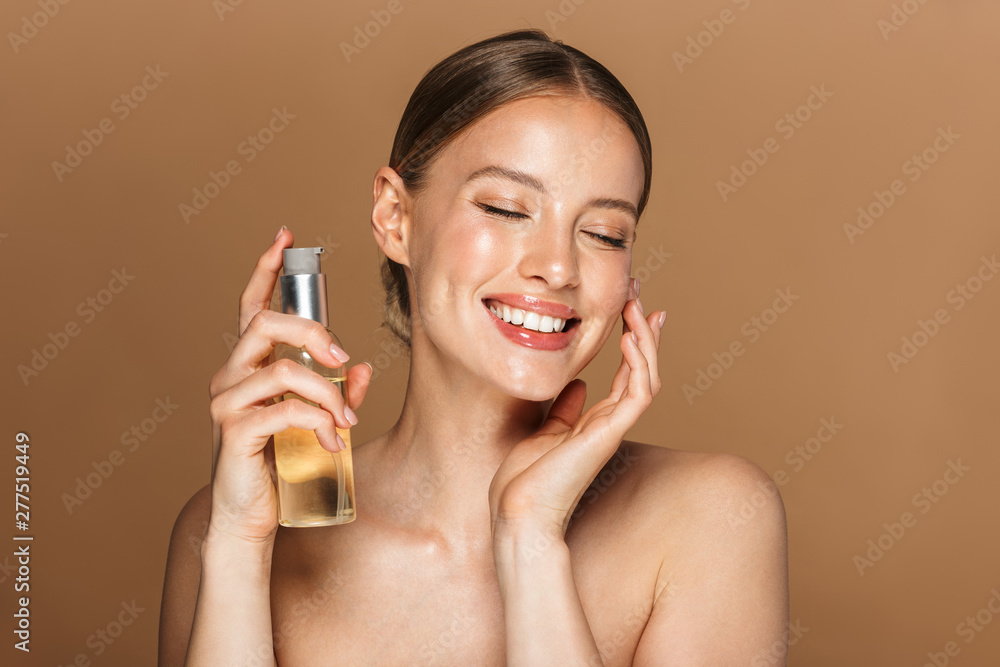 Image of adorable half-naked woman smiling at camera and holding face oil - obrazy, fototapety, plakaty 
