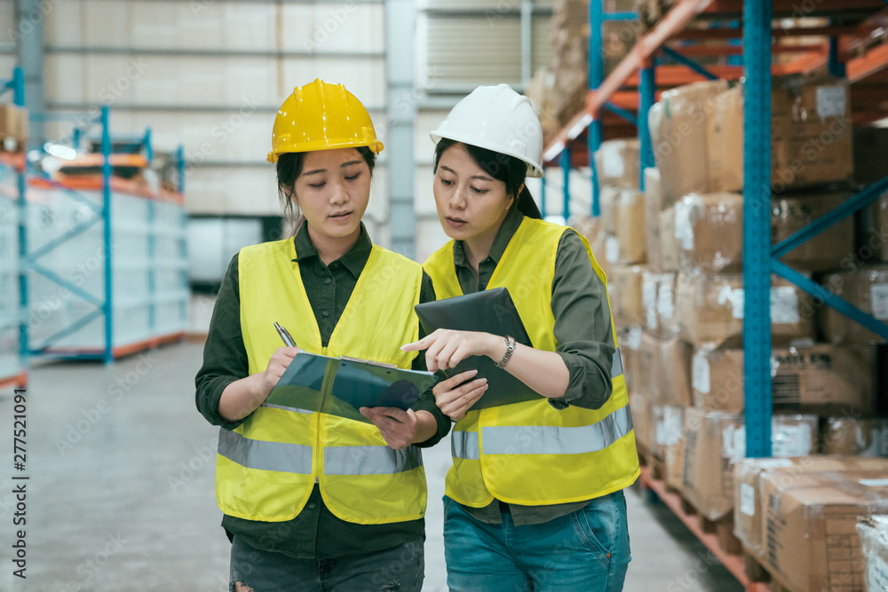 storehouse manager talking with worker in large warehouse. elegant female supervisor teaching new in employee girl while writing on clipboard document. two ladies staff in stockroom wear hard hats. storehouse manager talking with worker in large warehouse. elegant female supervisor teaching new in employee girl while writing on clipboard document. two ladies staff in stockroom wear hard hats.