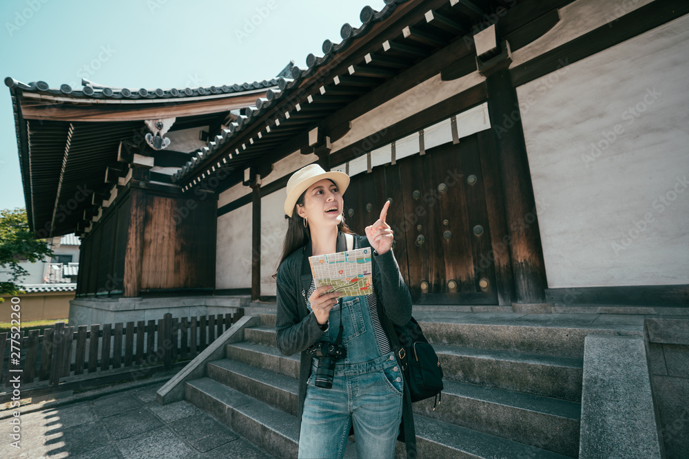 Happy Asian female tourist backpacker looking for direction while traveling around temple on summer holidays in osaka japan. girl traveler with camera holding paper map and point aside smiling.