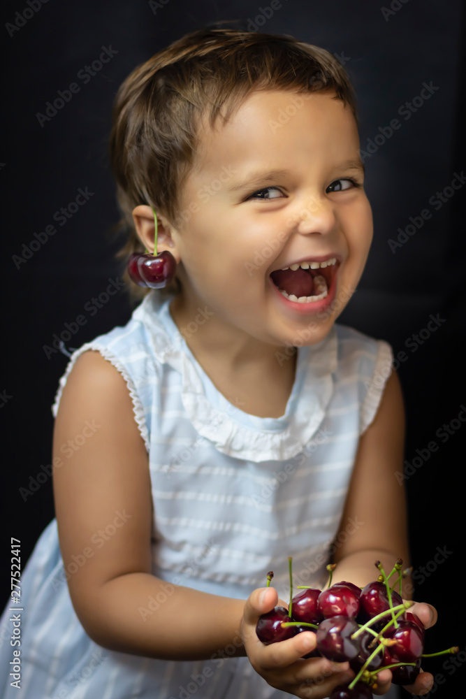 Cheerful little girl on a dark background holding cherries. Open space, the concept of a happy childhood, a happy child. Сoncept of organic food and healthy lifestyle, healthy food, happiness and joy Cheerful little girl on a dark background holding cherries. Open space, the concept of a happy childhood, a happy child. Сoncept of organic food and healthy lifestyle, healthy food, happiness and joy