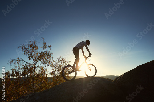 Silhouette of male cyclist riding on trial bicycle on top of big boulder, biker making acrobatic stunt on summer evening, blue sky and sunset on background. Concept of extreme sport outdoors