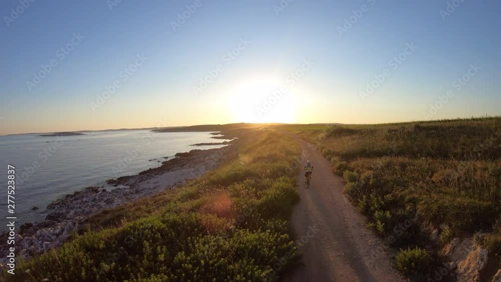 AERIAL Mountain biker riding his bike on a dusty road alojng the sea towards the sunset AERIAL Mountain biker riding his bike on a dusty road alojng the sea towards the sunset