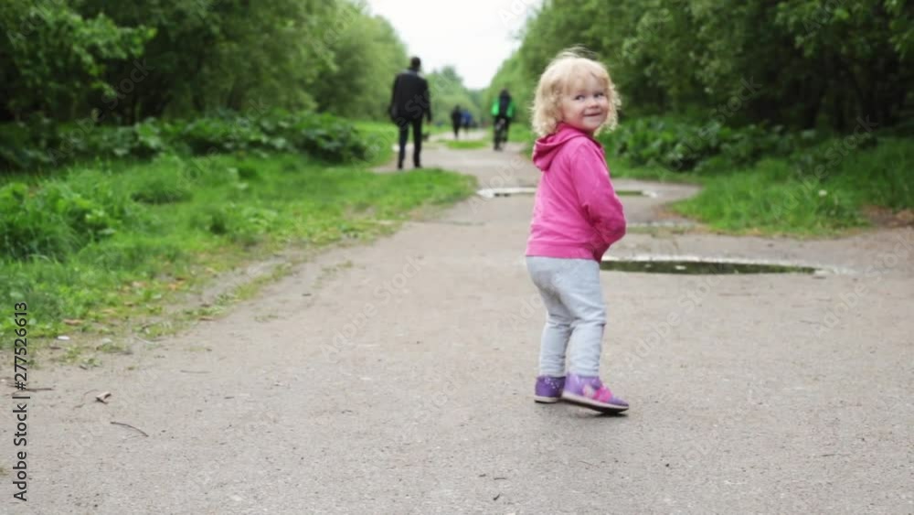 A happy girl is playing on the road after rain in a puddle she has fun. child looks at parents and comes in a puddle A happy girl is playing on the road after rain in a puddle she has fun. child looks at parents and comes in a puddle